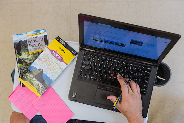 Overhead view of someone on a laptop with a map of Peru and a Guidebook to Machu Picchu beside it on the table.